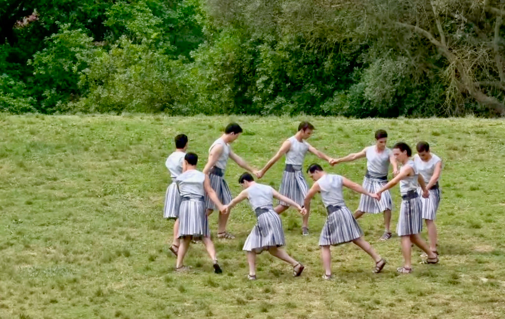 Male performers wear white and grey knee length skirts, holding hands in a circle, wide shot.