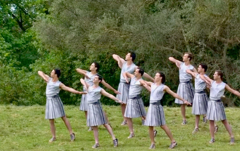 Male performers wear white and grey knee length skirts, on a grassy hill, with hands pointed backwards, and one elbow in air.