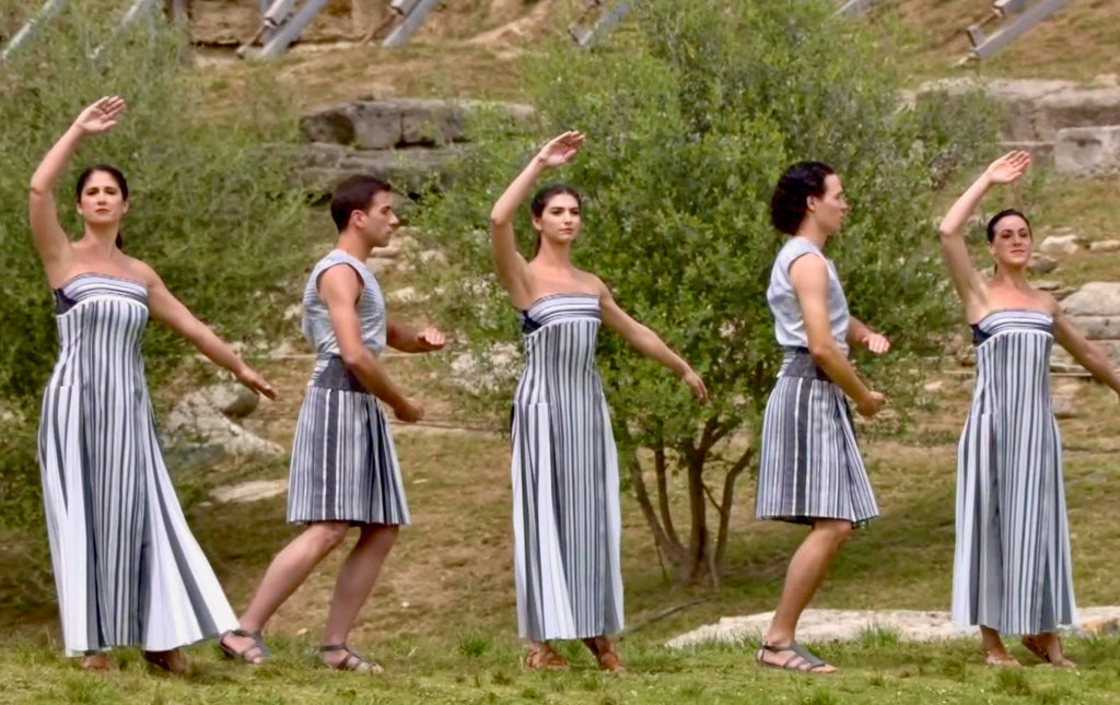 Male and female performers in white and grey vertically striped clothing, men wearing skirts, and woman wearing long dresses, alternating male/female.