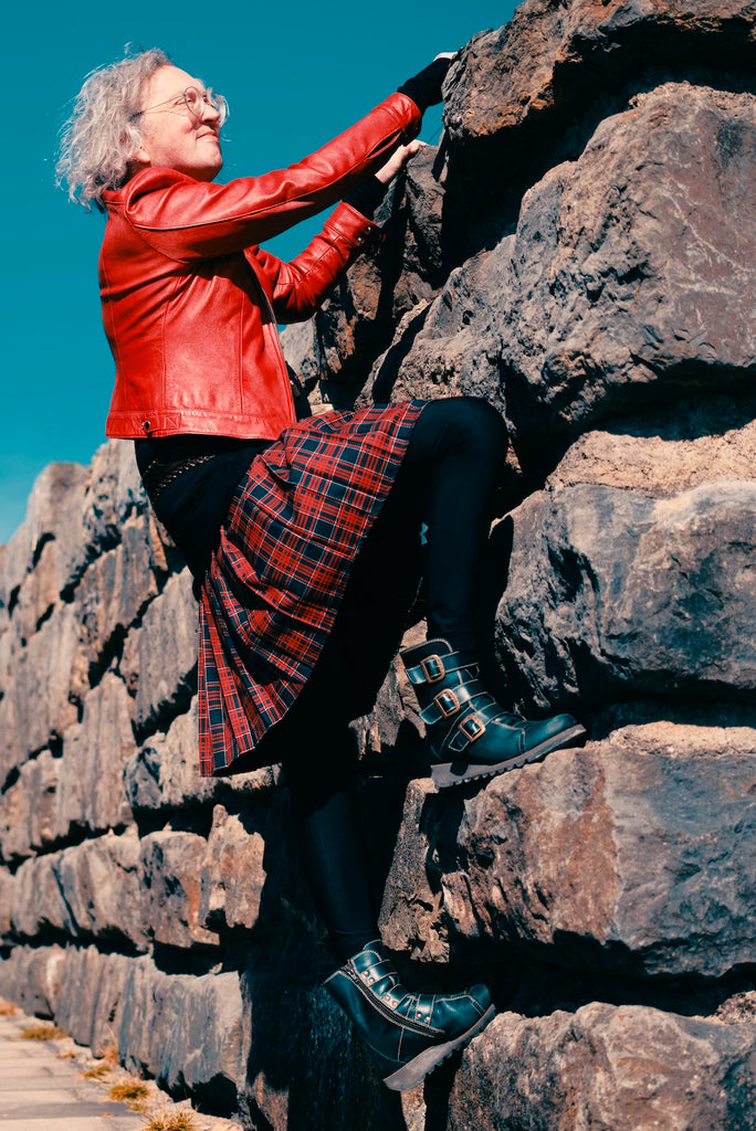 Andy climbing on a rock wall, peering over edge, with bright red leather jacket, red kilt-like skirt, and black boots.