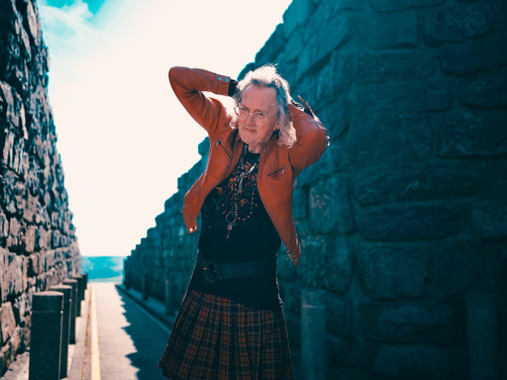 Andy holding his hair, in a skirt and leather jacket, between two rock walls.