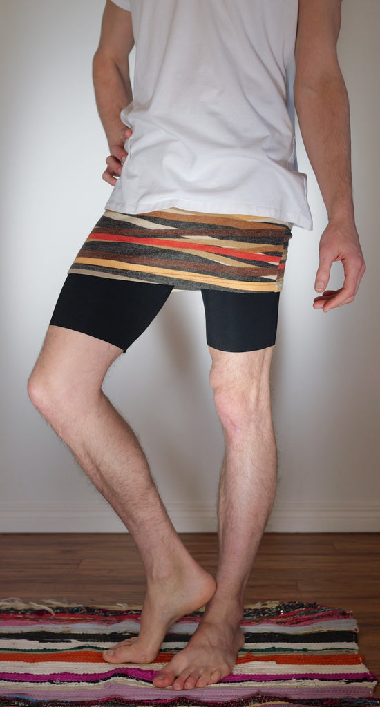Male bodied person wearing a long white tee that extends below waist, with orange/red/brown striped miniskirt, and long black underwear underneath.