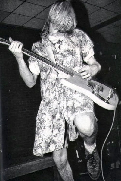 Kurt Cobain in a dress on stage, playing guitar, with one knee bent with that foot off the ground.