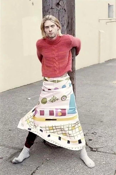 Kurt Cobain leaning against a telephone pole, on a sidewalk, wearing a women's red sweater and a colourful patterned long skirt, feet wide apart.