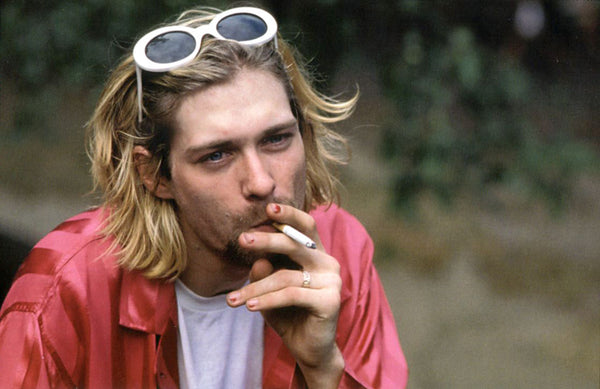 Kurt Cobain smoking in a red shirt, with white oval sunglasses at top of head, and wearing worn nail polish.