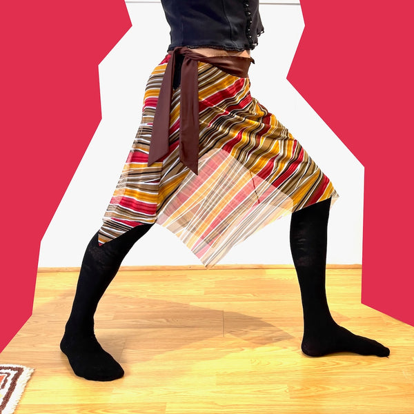 Me in orange and red striped mesh skirt, with brown ties hanging down. Black tights underneath, and feminine top (black). Posing in a stance.