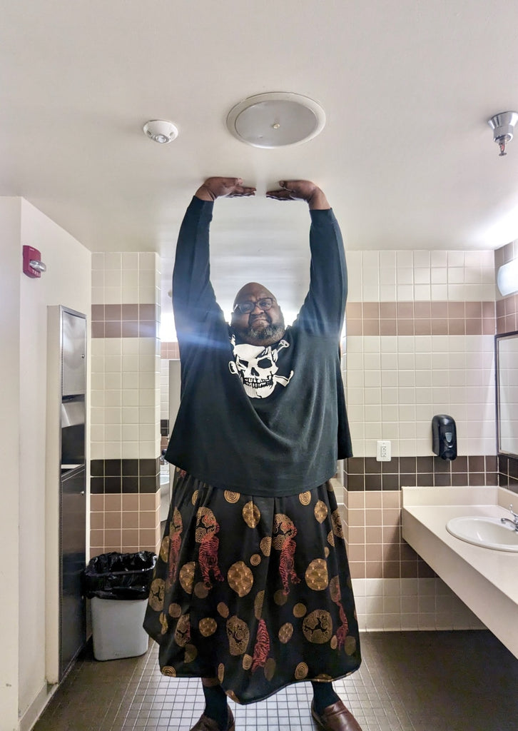 Phil, in a bathroom with hands pushing on ceiling, wearing a long black skirt with gold and orange tiger print, and a black long sleeve shirt with skull and cross bones.