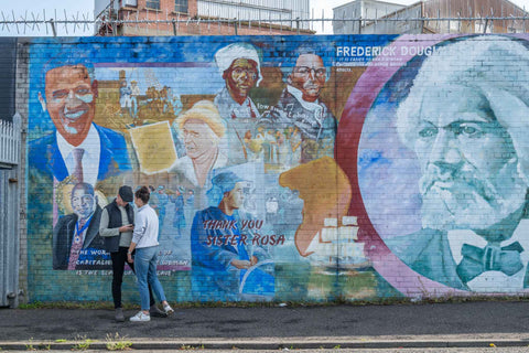 This photo was taken in Belfast, Northern Ireland. It is a brick wall that is painted with different colors of blue with a story about Frederick Douglas. There is a man and woman conversing in front of the wall.