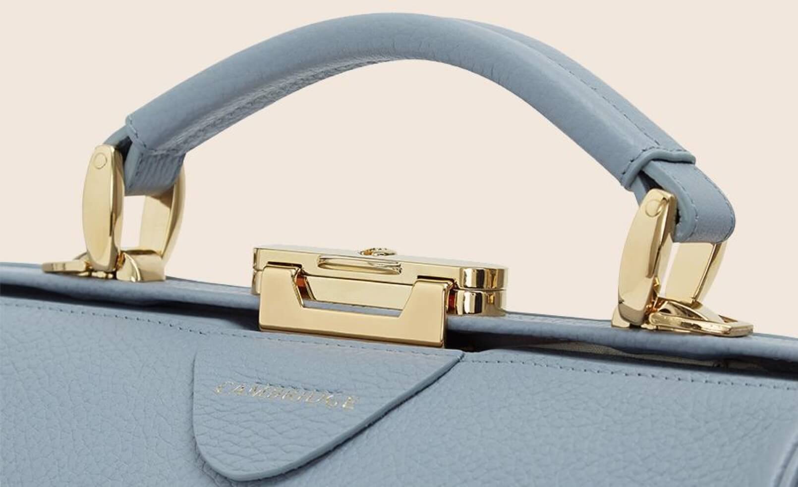 A closeup of a sky blue satchel handle with gold buckles. The leather detail and stitching is stunning.