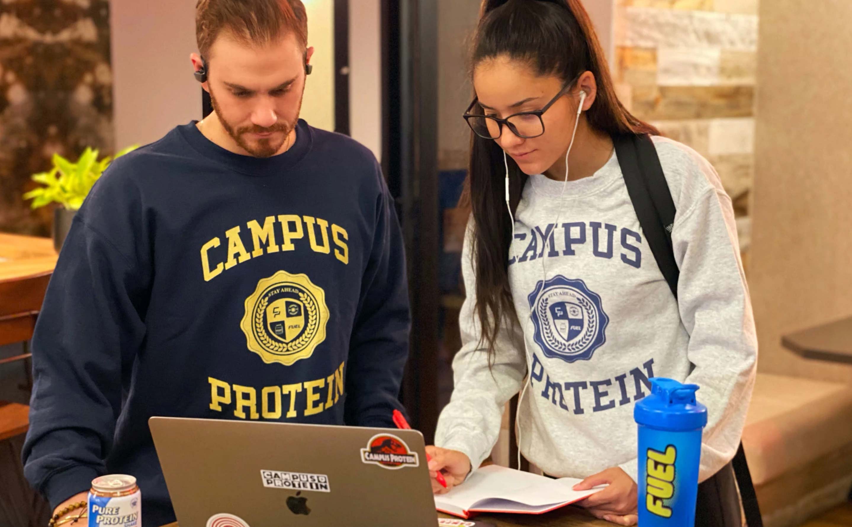 A male and a female student wearing campus protein crew neck sweaters in navy blue and grey, looking at a computer screen together.