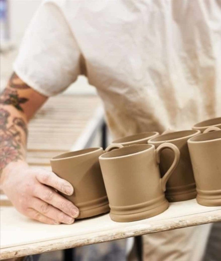 A ceramics artisan carefully unloads unfinished mugs from a wooden tray.