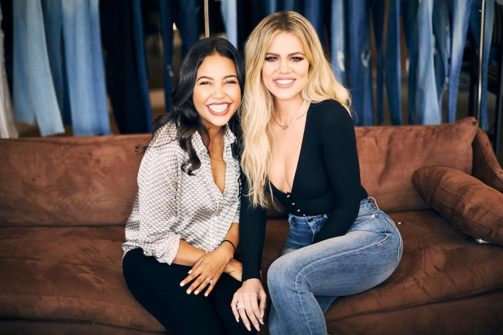 Good American co-founders Emma Grede and Khloe Kardashian smile as they sit on a fabric sofa.