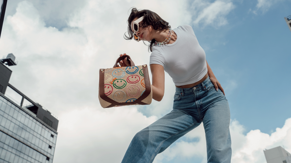 A woman holding a Smiley x WEAT tote bag with a blue sky background.