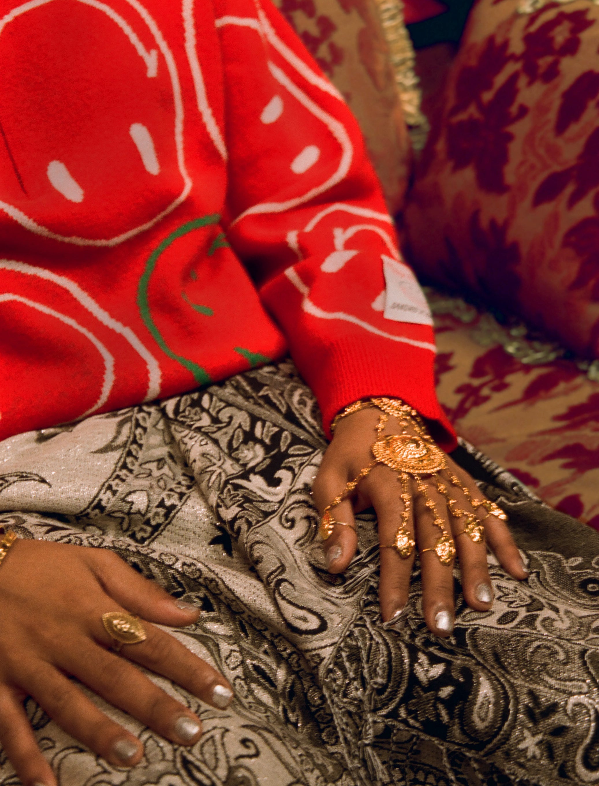 A hand covered in gold jewellery which and a body wearing a red jumper from Sandro & Smiley. 