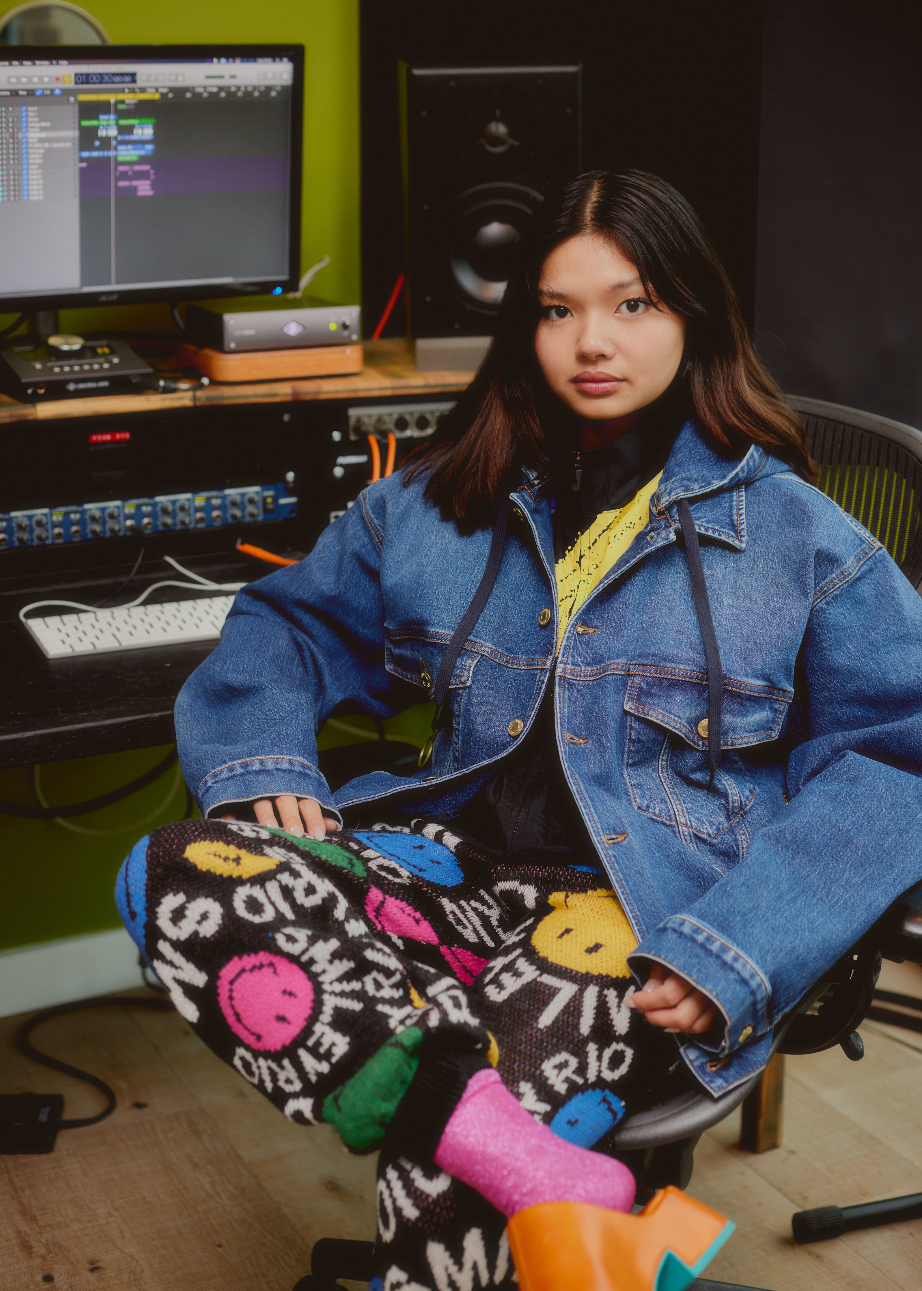 Lucy Tun wearing a Reebok x Smiley jacket and a Farm Rio x Smiley pants and looking at the camera whilst sitting on a chair.