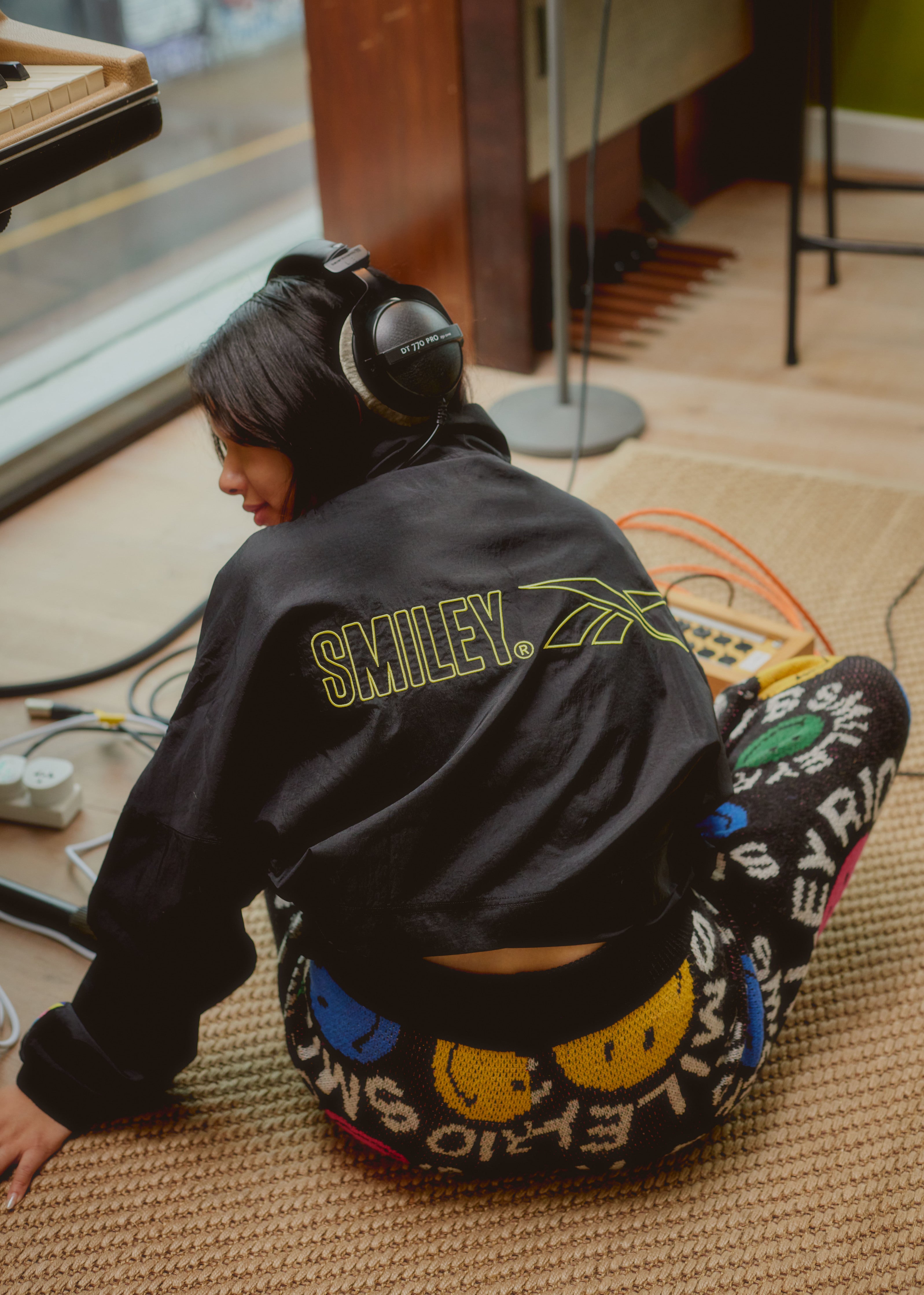 Lucy Tun wearing a Reebok x Smiley jacket and Farm Rio x Smiley pants whilst sitting on the floor with her back to the camera.