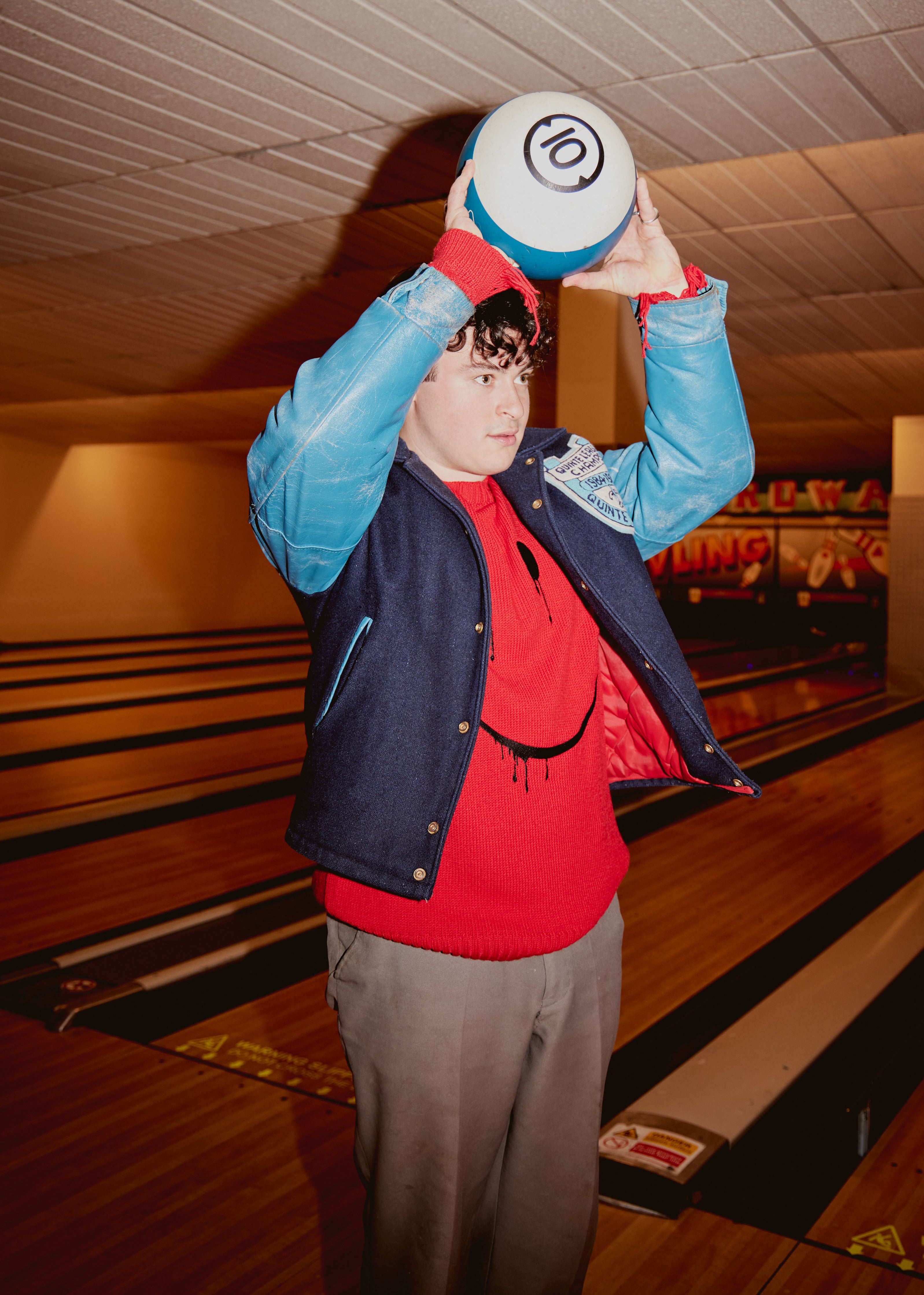 Dan from KAWALA is wearing a Stuff Maker x Smiley embroidered sweater holding a bowling ball above his head.