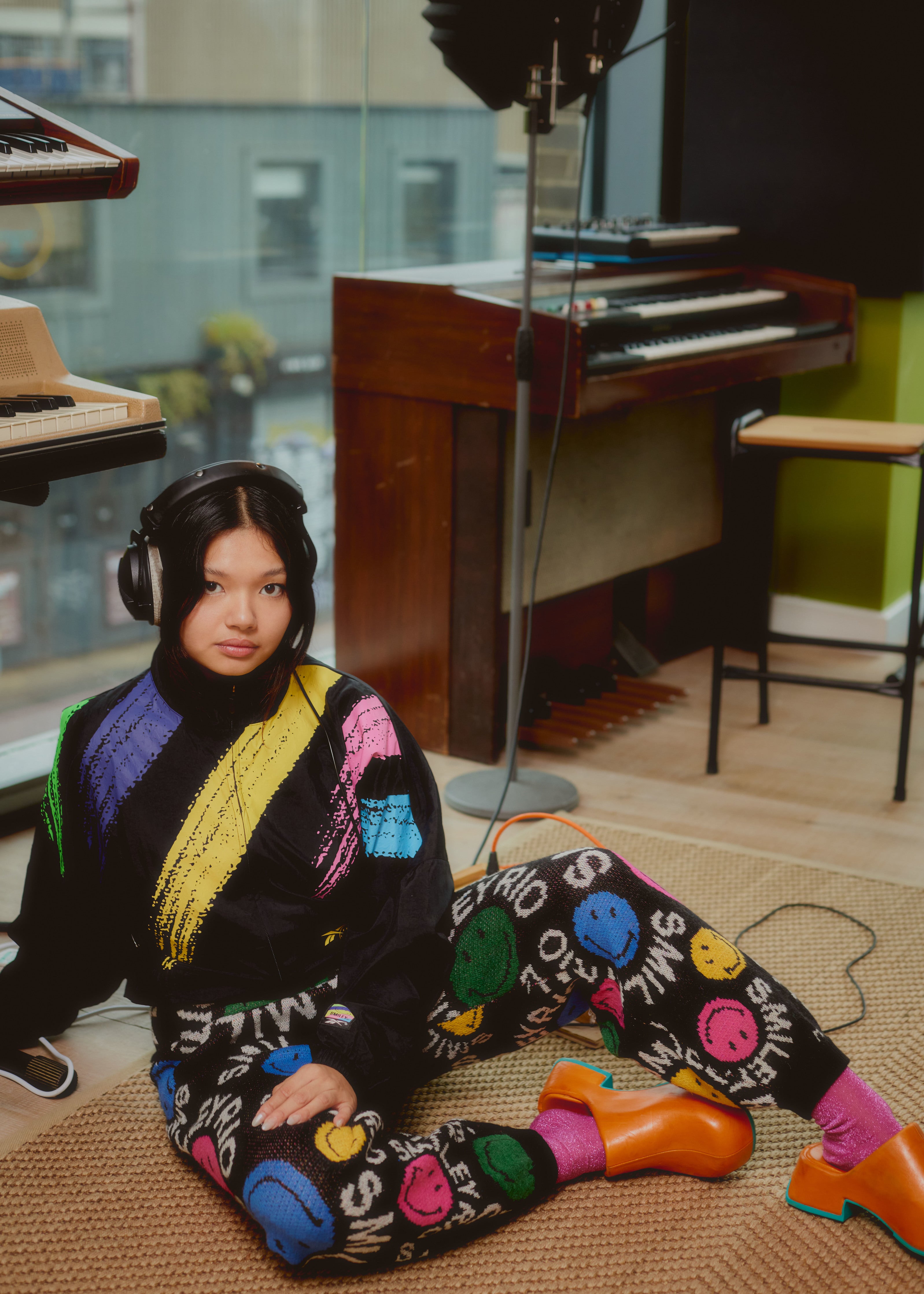 Lucy Tun wearing a Reebok x Smiley jacket and Farm Rio x Smiley pants whilst sitting on the floor looking up at the camera.