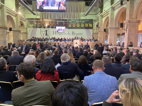 A sea of people seated in a hall before a stage draped with the Hospices de Beaune logo with auctioneers on stage