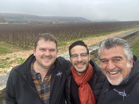 Matt, Marco, and Roberto s miling at the camera in front of a vineyard in foddy weather with a low range of hills in the background