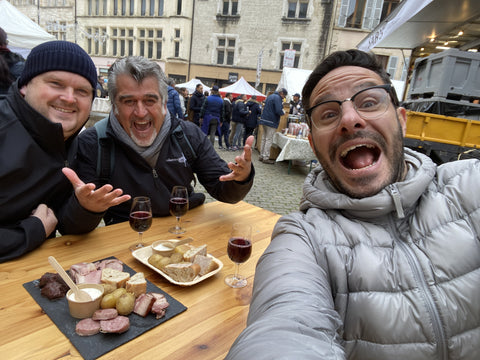 Matt, Roberto Cipresso, and Marco mugging at the camera at a wooden table covered with wine and small plates on a cobblestone street with crowds and vendor tents in the background