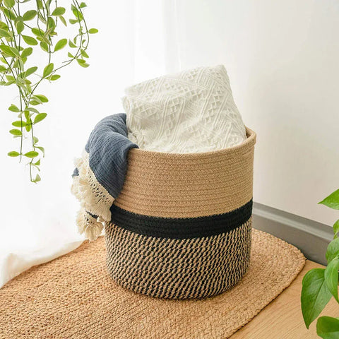 The image shows a round woven jute basket. The bottom portion of the basket features a black and natural fiber herringbone pattern, while the top portion is a solid natural jute color. The basket contains a folded white textured blanket and a partially visible blue textured blanket with fringe. The basket sits on a natural fiber rug or mat on a light-colored wooden floor. Green leafy plants are visible in the periphery, suggesting an indoor setting near a window with natural light.