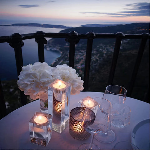 Four lit candle holders of varying styles and heights illuminate a round table covered with a white tablecloth on a balcony overlooking a scenic coastal view at dusk or dawn. White floral arrangements and several empty wine glasses are also on the table. A black wrought-iron railing borders the balcony, with the sea and distant land visible in the background under a cloudy sky.