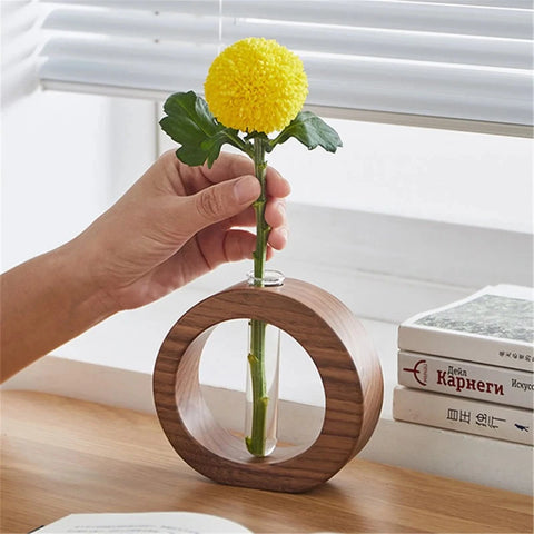 A hand places a stem with a round, yellow chrysanthemum into a clear glass test tube held within a circular wooden vase. The vase sits on a wooden desk near a window with white blinds and a stack of books.
