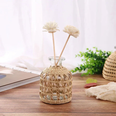 A small, cylindrical glass vase partially covered in woven rattan sits on a wooden surface. The vase holds two artificial white flowers with thin wooden stems. A blurred background shows a window with white curtains, a book, and some greenery.