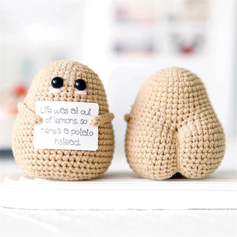Two small, light brown crocheted potato-shaped figures stand on a white surface. The figure on the left faces forward with small black eyes and holds a white tag that reads, "Life was all out of lemons, so here's a potato instead." The figure on the right faces away, showing its crocheted texture. A blurred background is visible.
