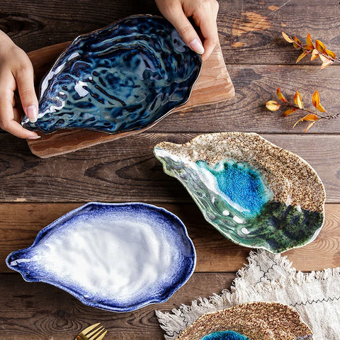 Three uniquely glazed ceramic oyster dishes in blue, white, and brown tones, arranged on a wooden board and table with decorative elements.