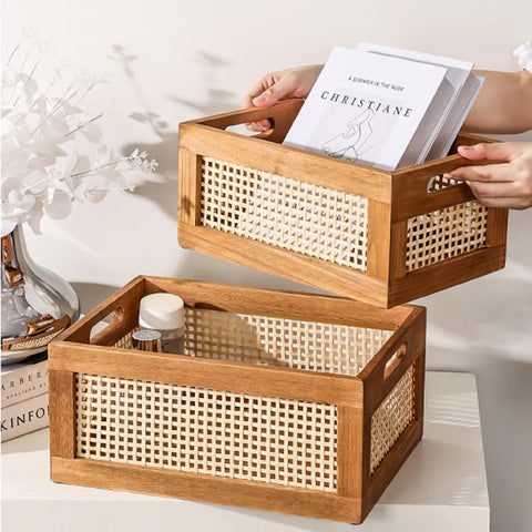 Two rectangular wooden storage baskets with woven rattan side panels sit on a white surface. The top basket is being held by two hands and contains a stack of white books or papers, one with the title "CHRISTIANE" visible. The bottom basket contains a clear glass bottle with a white lid and other small items. Both baskets have cutout handles. A blurred background shows a white wall and a glass vase.