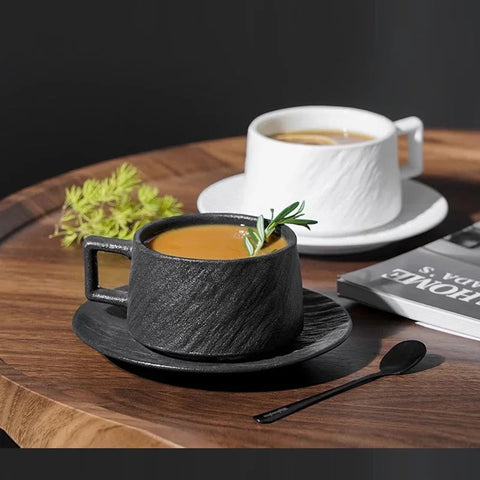 A black and white ceramic coffee cup set with a textured rock-like finish, placed on matching saucers on a wooden table. The black cup is filled with coffee and garnished with a sprig of rosemary, while the white cup holds a beverage in the background. A black spoon and a magazine are also on the table, adding to the modern, minimalist aesthetic.
