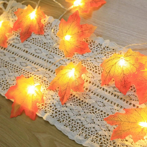 A string of warm white fairy lights shaped like autumn maple leaves is arranged on a delicate white lace runner. The artificial leaves are orange and red with visible veins, and the lights within them emit a soft, inviting glow. The lace runner has an intricate pattern of small openings.