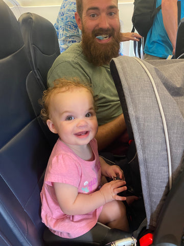a toddler sits in her carseat on a flight, smiling at the camera, next to her father