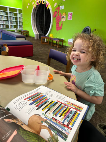 a toddler-aged girl sits at a library table, eating with a pink and orange bento box