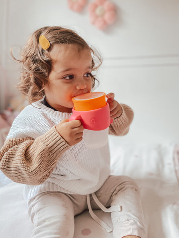 a young toddler drinks from a pink and orange 360 cup