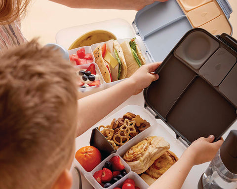 a boy looks down at a bento box that includes a pastry, pretzels, and fruit