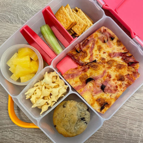 a b.box lunchbox containing flatbread pizza, crackers, fruit, and a muffin