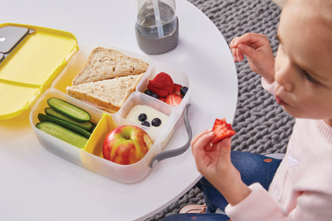 a child eats from a yellow b.box lunchbox containing a sandwich, yougurt, an apple, berries, and cucumber slices