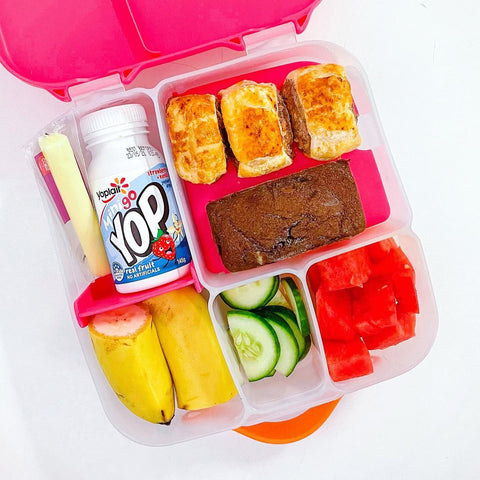 an open bento box featuring sandwiches, a brownie, a cheese stick, yogurt, a banana, and vegetables