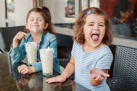 Two funny Caucasian little preschool sisters siblings drink milk shakes in a cafe. Friends girls having fun together. Cold summer desserts for kids. Happy authentic childhood lifestyle