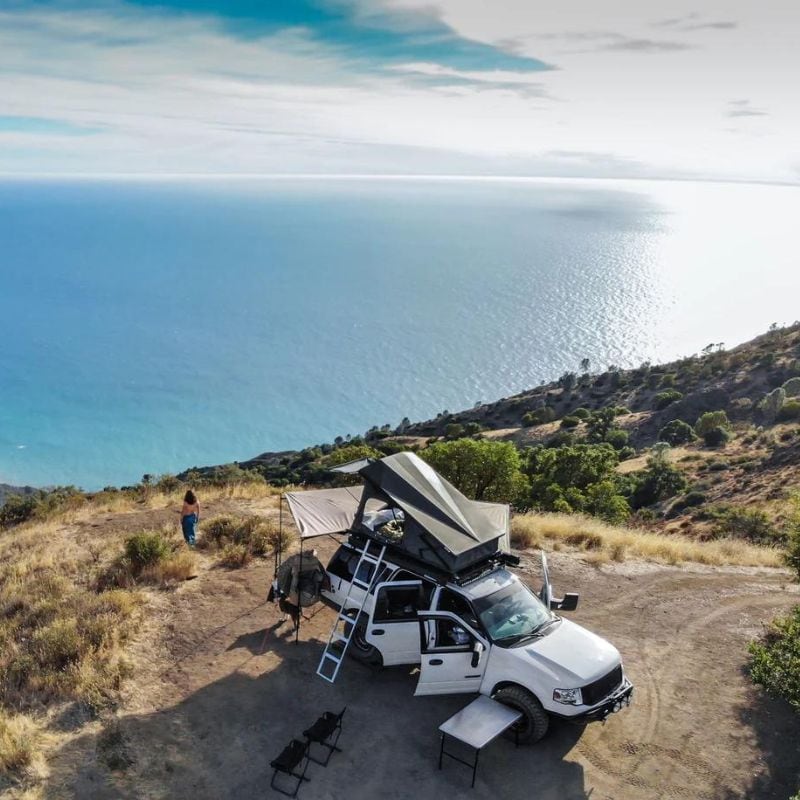 eezi-awn-stealth-hard-shell-roof-top-tent-open-drone-view-on-ford-explorer-with-person-outside-camping-next-to-the-ocean