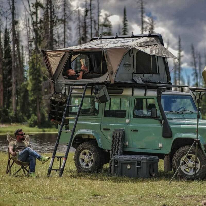 overland-vehicle-systems-bushveld-hard-shell-roof-top-tent-open-front-corner-view-on-land-rover-defender-in-green-field-with-persons-camping
