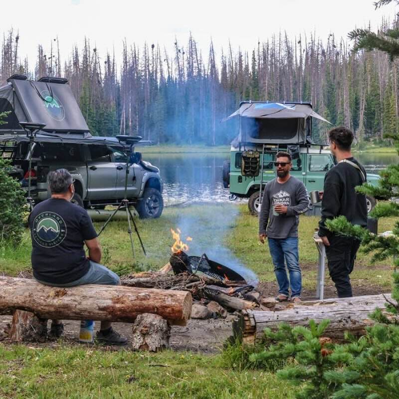 overland-vehicle-systems-bushveld-hard-shell-roof-top-tent-open-side-view-on-land-rover-defender-in-forest-with-people-at-the-campfire