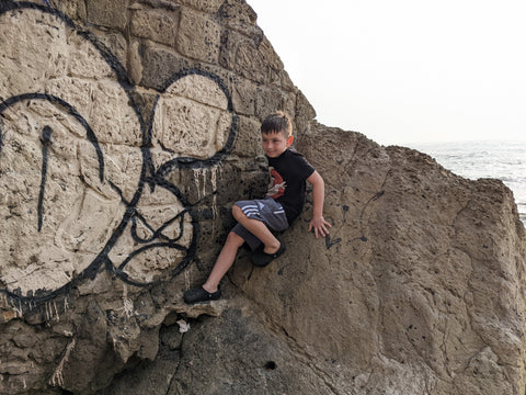 Young boy leaning against a rock with graffiti art on it. The sea can vaguely be seen in the background.