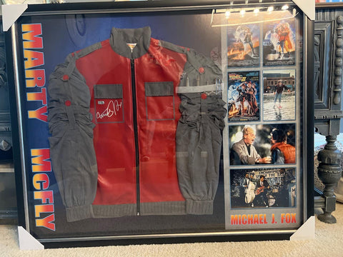 Image of a high-quality replica of the 'Back to the Future' jacket, adorned with Michael J. Fox's signature. The vibrant red and grey jacket, symbolic of the iconic 80s film, is displayed in a protective case, preserving the autograph and the jacket's immaculate condition. A personalized note of gratitude from a delighted customer, Teri, adds a warm, personal touch to the image