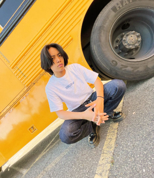 Man wearing Pursuit of Knowledge Shirt crouched down in front of a yellow school bus.