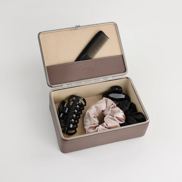 An open, rectangular jewelry box made of brown leather with beige lining is visible. Inside there is a black comb, a hair clip and two hair ties. This box offers an elegant and safe storage solution for jewelry and accessories and is equipped with a compartment in the lid for smaller items. The lettering "nimbus" and the logo "F. Hammann - Made in Germany" are visible.