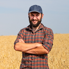 a man in a flannel shirt and denim baseball cap standing in a wheat field with his arms folded